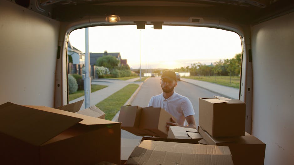 A man with a beard and glasses, wearing a white polo shirt and cap, standing outside a moving van during daylight, loading or unloading multiple brown cardboard boxes and smaller parcels. The van's re