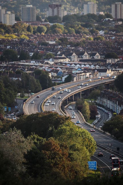 Aerial view of a multi-lane curved road with moving cars and trucks, situated in an urban area surrounded by green trees and residential buildings. The road appears to be part of a home relocation route, with a bridge crossing over other roads below. In the background, there are densely packed houses, commercial buildings, and high-rise apartments, indicating a busy cityscape. The weather is clear, with natural daylight illuminating the scene. The image captures the practical aspects of transport logistics, relevant to furniture transport and packing during house removals, as performed by companies like Man and a Van Enfield as they prepare for efficient home relocation and moving services.