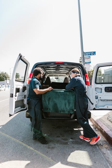 Two movers from Man and a Van Enfield are loading a large piece of furniture, covered with a green blanket, into the back of a black panel van parked on a street. The van's rear doors are wide open, revealing a spacious interior with additional packing materials. One mover, wearing a dark uniform, is inside the van passing the furniture to another worker standing outside, dressed in a blue t-shirt and black trousers, both focused on the lifting and positioning process. The scene takes place during daylight, with clear weather and a street sign visible in the background indicating nearby locations. This image illustrates the household packing and furniture transport phase within a home relocation or moving service, highlighting careful handling and logistical coordination involved in house removals, as offered by Man and a Van Enfield.