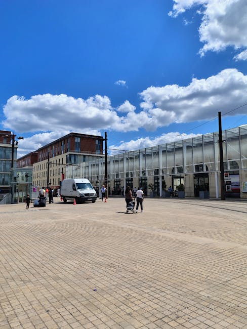 A spacious outdoor paved area in front of a modern building with large glass windows and a metallic facade, under a bright blue sky with scattered white clouds. Several people are visible, some standing near a white van parked on the open space and others walking, with some carrying or preparing large cardboard boxes and wrapped furniture for removal. The scene suggests a home relocation or furniture transport process, supported by the presence of removal equipment like trolleys and blankets, with the building likely serving as a station or commercial property. The setting is well-lit with natural daylight, and the environment appears organized for moving activities, aligning with services offered by Man and a Van Enfield for house removals and moving logistics.