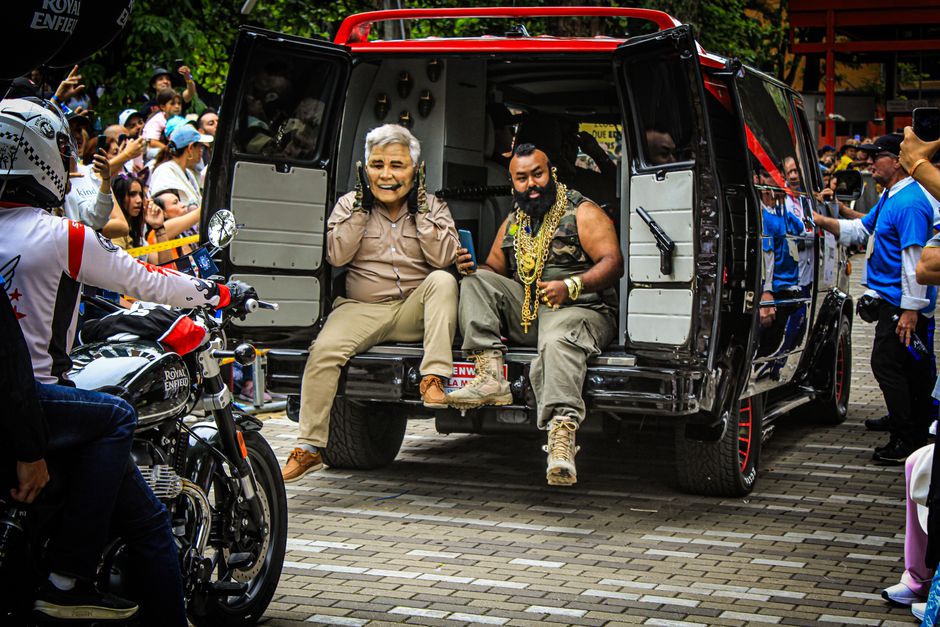 A woman with short grey hair, dressed in a beige shirt and light-colored trousers, sitting on the edge of a black vehicle's low tailgate, smiling and talking on a mobile phone. Beside her, a man with a beard, wearing a sleeveless camouflage shirt, grey cargo pants, and beige combat boots, is seated with one arm resting on his knee, holding a smartphone in the other hand. The vehicle, likely a van used for furniture transport or home relocation, has its rear doors open, revealing the empty interior with visible padding and structural supports. Both individuals are partly inside the vehicle, positioned on the tailgate, which is lowered onto a paved surface made of grey bricks. Surrounding them are several people, some in casual clothing, observing or taking photos, with children and adults visible in the background. A police officer on a Royal Enfield motorcycle is approaching, and other vehicles and banners can be seen in the background of what appears to be a public event or outdoor gathering related to moving or community activities. The scene reflects a moving or transport setting, with the vehicle prepared for loading or unloading furniture or equipment as part of a logistics process, and the environment is well-lit with natural daylight.