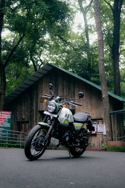 A black and grey electric motorcycle with green accents is parked on a paved surface in front of a wooden cabin in a forested area. The motorcycle features a round headlight, rearview mirrors, and a comfortable seat, with a small storage compartment attached to the side. The background consists of tall trees with green foliage and sunlight filtering through the branches, creating a natural canopy. To the right of the motorcycle, a metal railing is visible, and the wooden cabin has a sloped green roof, wooden siding, and a notice board on the wall. This scene depicts a quiet outdoor setting suitable for nature-based activities or home relocation logistics involving packing and transport, with the environment suggesting a peaceful woodland location, consistent with scenic areas like Forty Hall and Bullsmoor, ENFIELD. Man and a Van Enfield’s vehicle is not visible, but the setting reflects the kind of environment where moving and transportation services could be involved in a home relocation process.