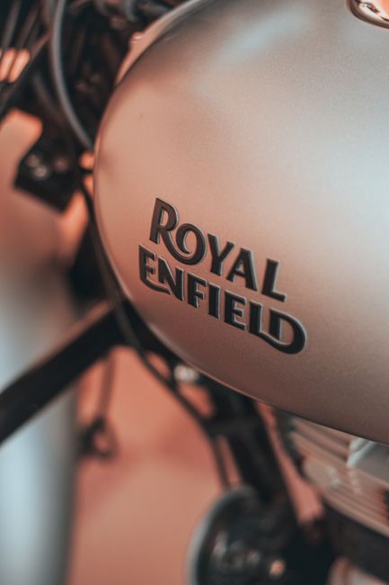 Close-up image of a metallic silver Royal Enfield motorcycle fuel tank with the brand logo clearly visible, situated inside a property with soft lighting. In the background, part of the handlebars, front wheel, and some packaging materials such as cardboard boxes and plastic wrapping are visible, indicating a home relocation process. The motorcycle is positioned near a doorway or doorway area, suggesting it is being prepared for transport or loading onto a vehicle. The scene includes a trolley and straps used for securing the motorcycle during transport, reflecting aspects of furniture or vehicle transport involved in house removals services. The environment appears organized, with a focus on safely handling valuable items during a move, aligning with professional moving and packing procedures offered by Man and a Van Enfield, as part of their house removals services in the Forty Hall and Bullsmoor area.